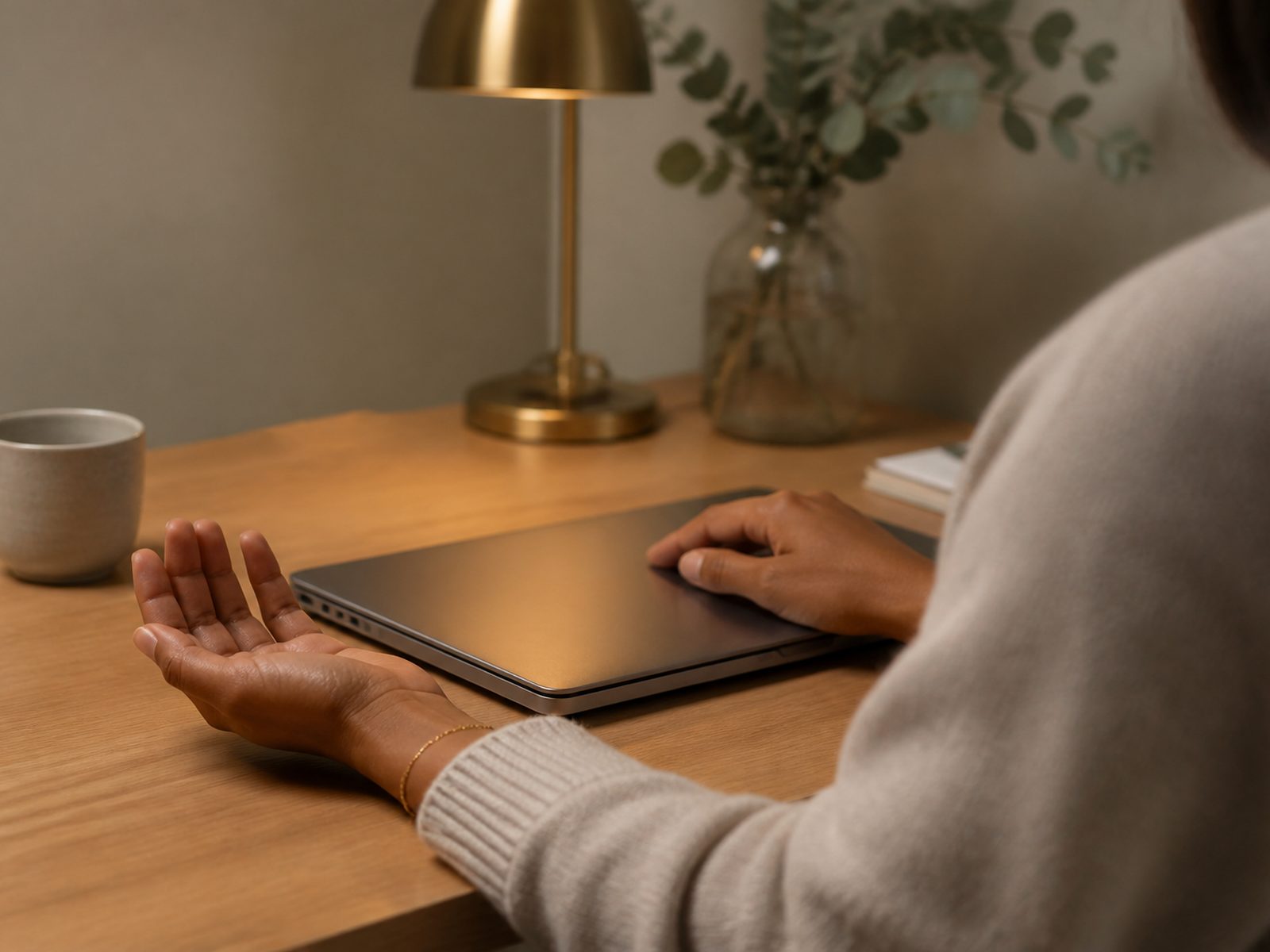 Same shoot, relaxed hands on a closed laptop at an oak desk with lamp and eucalyptus.